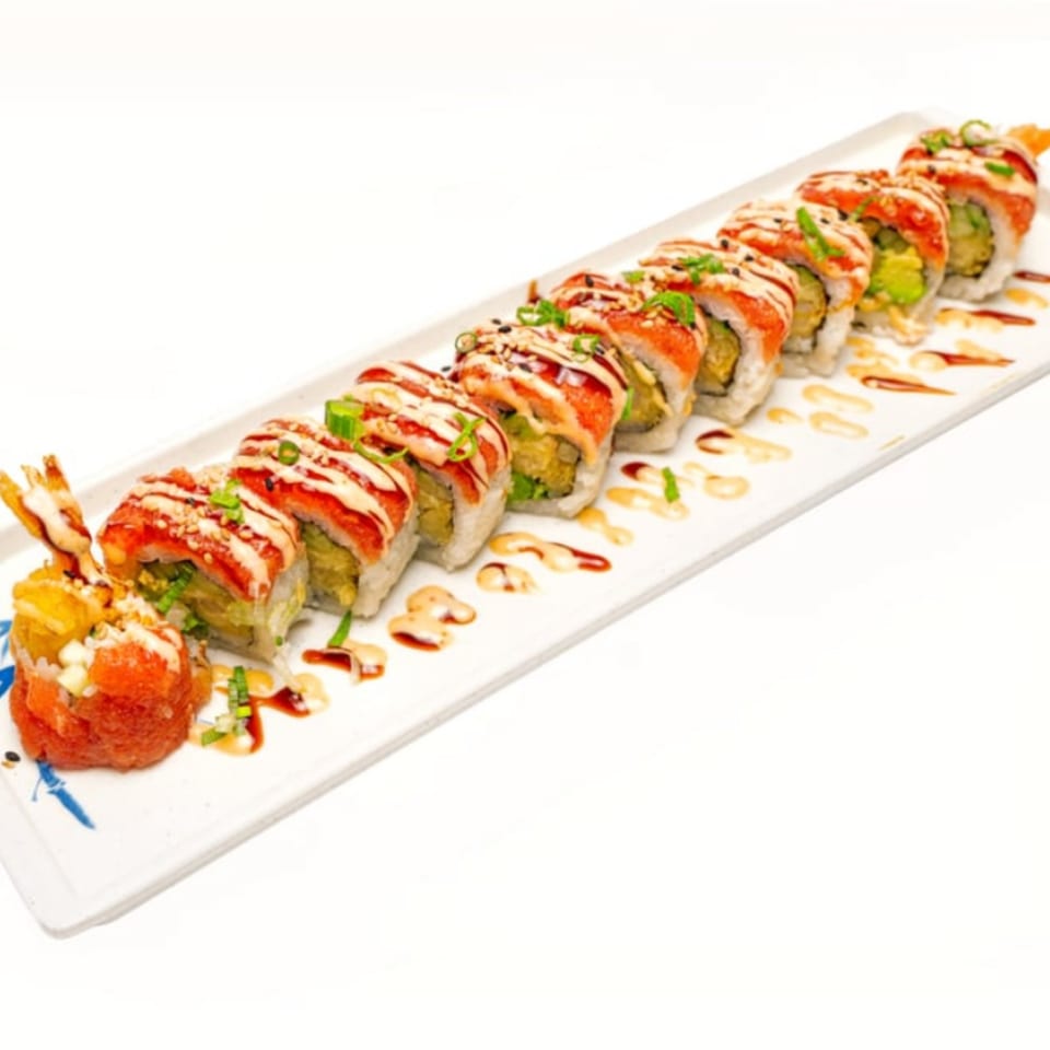 A long white plate holds a row of sushi rolls topped with red fish, sauces, and green onions.